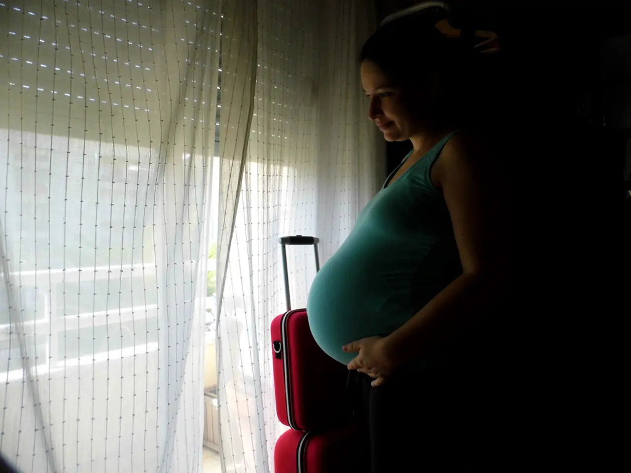 There is a pregnant lady standing in front of a white curtain and there are red luggage bags beside...
