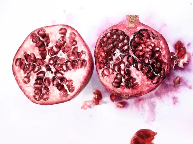 In the image there is a half cut pomegranate on a table with its seeds in front of it.